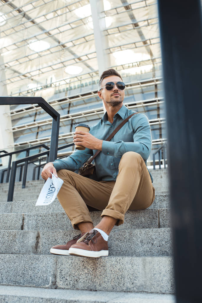 low angle view of man in sunglasses holding coffee to go and business newspaper while sitting on stairs - Photo, Image