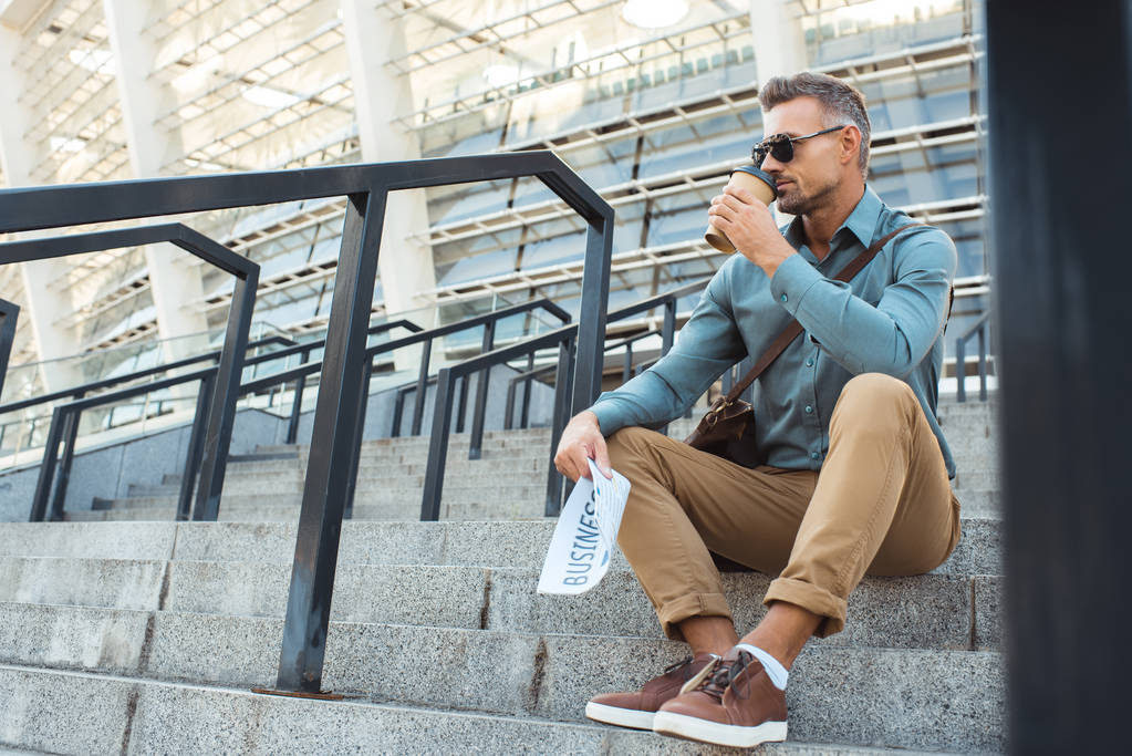 pohledný muž v sluneční brýle drží obchodních novin a pití kávy z papírového kelímku, zatímco sedí na schodech - Fotografie, Obrázek