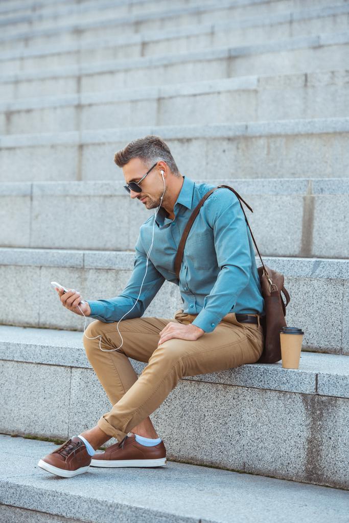 středního věku krasavec v sluneční brýle a sluchátka sedí na schodech a pomocí smartphonu - Fotografie, Obrázek