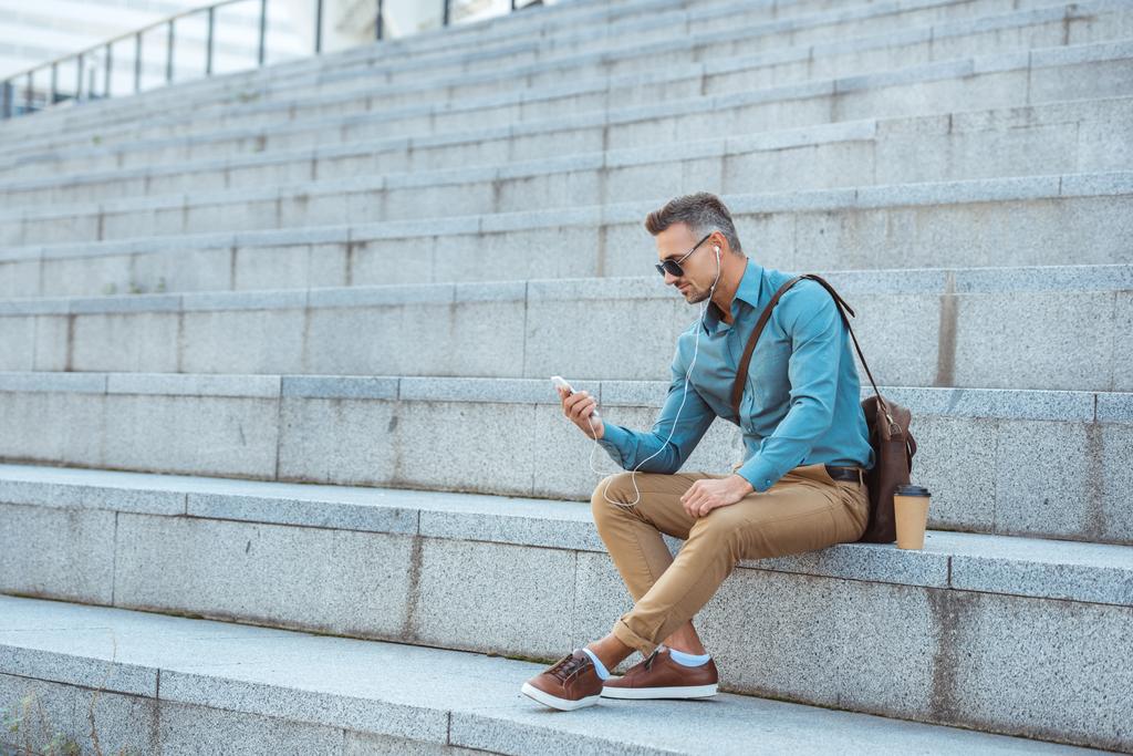 όμορφος μοντέρνος άνδρας στα ακουστικά και γυαλιά ηλίου που κάθονται στα σκαλοπάτια και χρησιμοποιώντας το smartphone   - Φωτογραφία, εικόνα