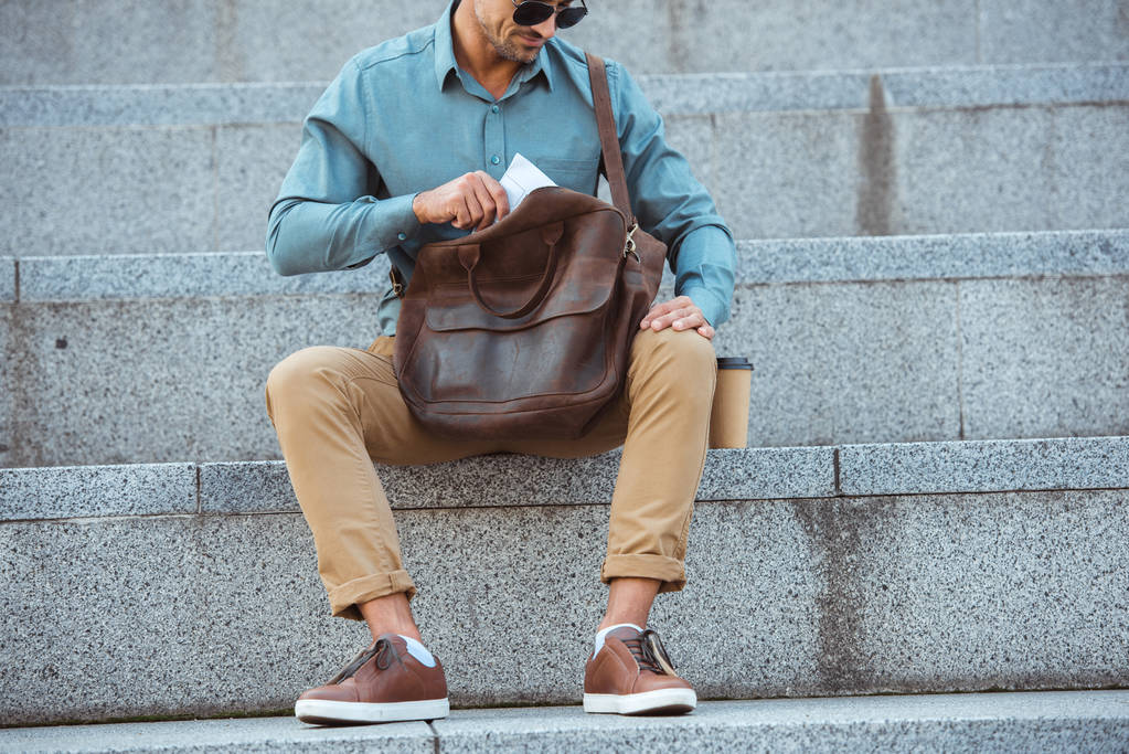 cropped shot of man in sunglasses sitting on stairs and putting newspaper into leather bag - Photo, Image