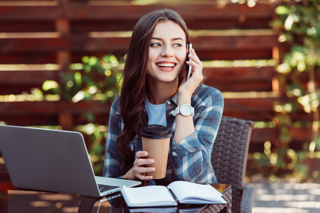 Lachende vrouw met koffie te gotalking op smartphone bij tafelblad met laptop en notebook - Foto, afbeelding