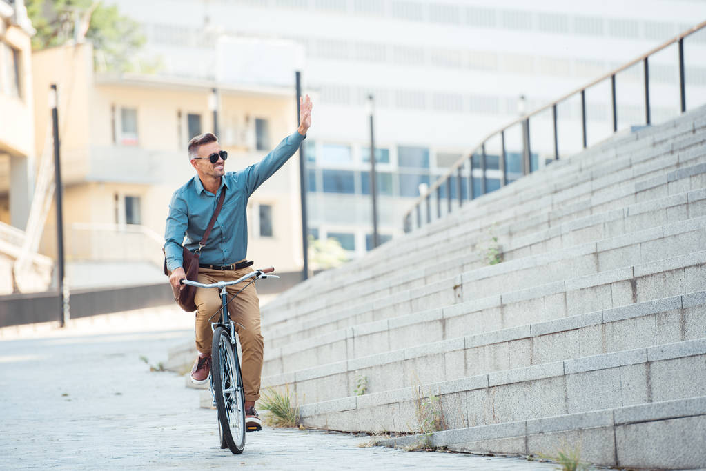 usmívající se muž v sluneční brýle jízda kole a mávání rukou na ulici - Fotografie, Obrázek