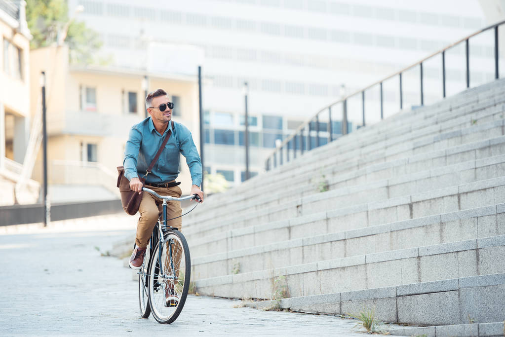 handsome middle aged businessman in sunglasses riding bike and looking away on street  - Photo, Image