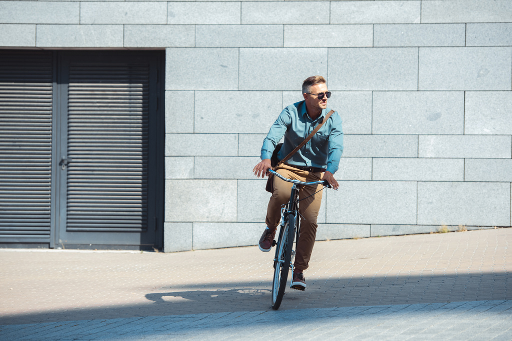κομψό μέσης ηλικίας άνδρα σε γυαλιά ηλίου ιππασία ποδήλατο και αναζητούν μακριά στην οδό  - Φωτογραφία, εικόνα