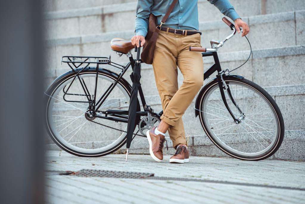 обрезанный снимок мужчины, держащего зонтик и опирающегося на велосипед на улице
 - Фото, изображение