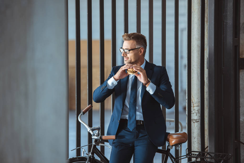 Lächelnder Geschäftsmann mittleren Alters mit Brille sitzt auf dem Fahrrad und hält Hamburger in der Hand  - Foto, Bild
