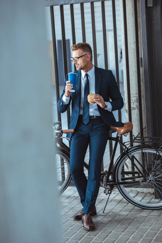 vista completa de hombre de negocios guapo sosteniendo hamburguesa y beber de la taza de papel mientras está sentado en bicicleta y mirando hacia otro lado
 - Foto, imagen