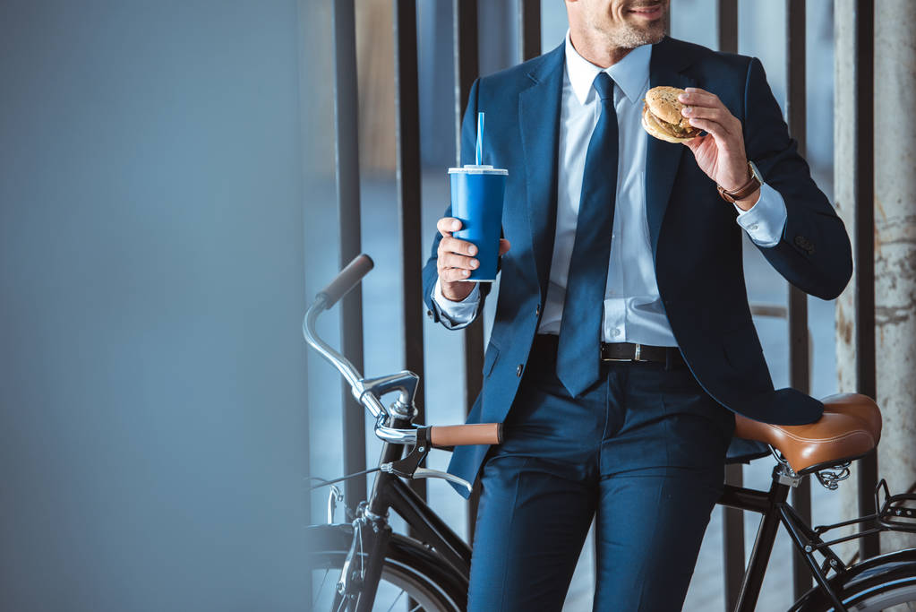 tiro recortado de empresário sorridente segurando copo descartável e hambúrguer enquanto sentado em bicicleta
 - Foto, Imagem