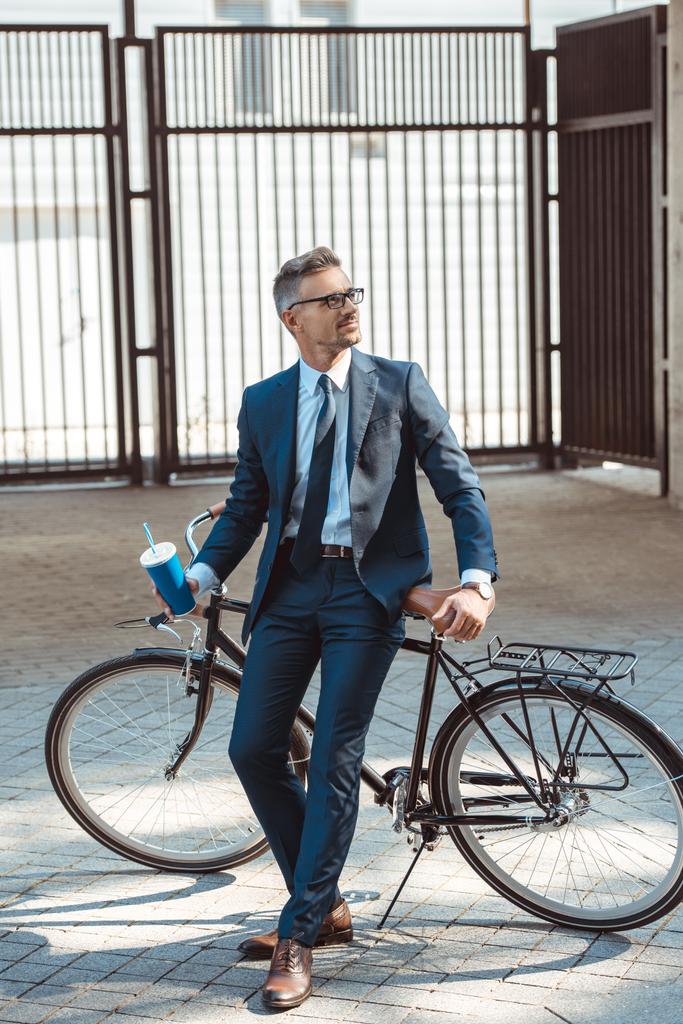 bonito sorridente empresário segurando copo de papel e sentado na bicicleta na rua
 - Foto, Imagem