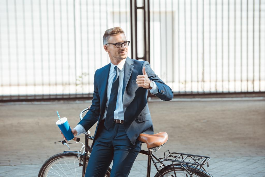smiling businessman sitting on bicycle, holding paper cup and showing thumb up on street - Photo, Image