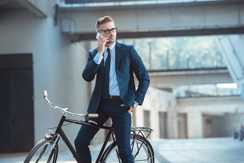 handsome businessman sitting on bike and talking by smartphone on street - Photo, Image
