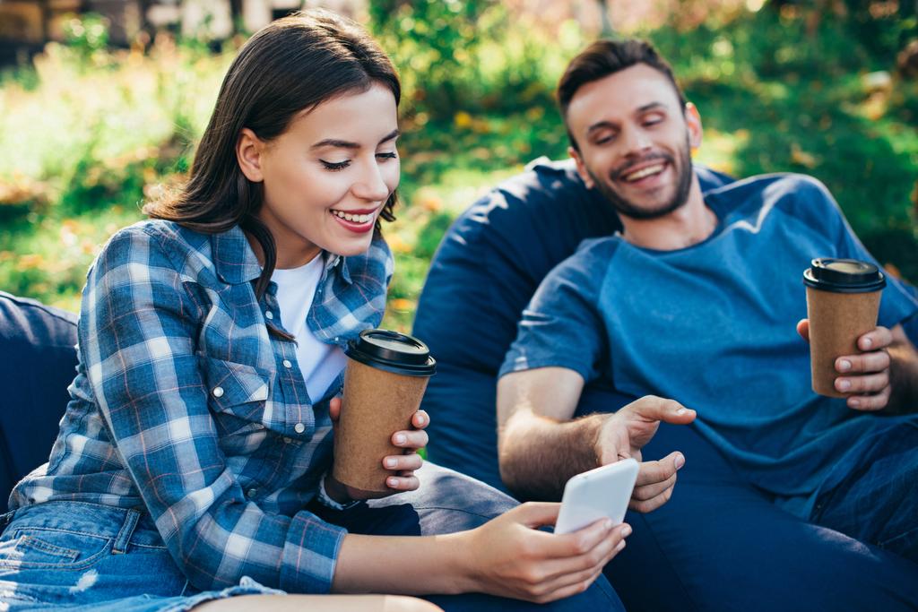 uśmiechnięty znajomych patrząc smartphone na krzesła worek fasoli w parku i gospodarstwa w papierowych kubków kawy - Zdjęcie, obraz