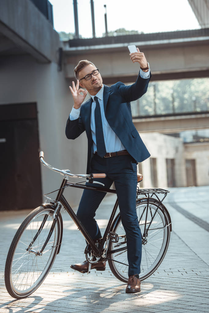 smiling businessman showing ok sign to smartphone while sitting on bicycle on street - Photo, Image