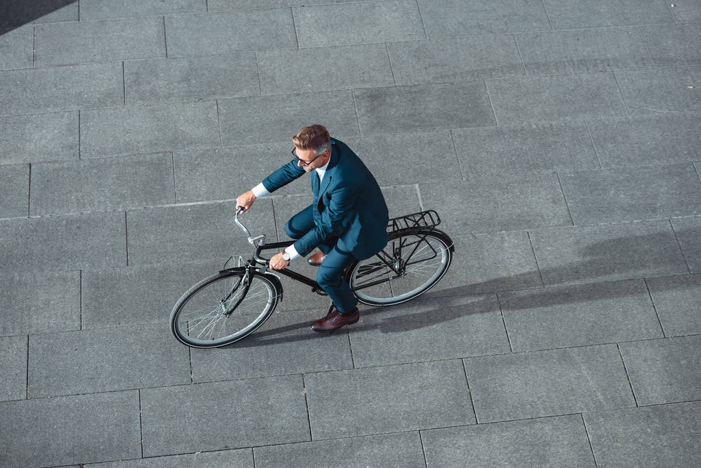 μεγάλη γωνία προβολής της μέσης ηλικίας επιχειρηματία σε κοστούμι και γυαλιά, ιππασία ποδήλατο στο δρόμο - Φωτογραφία, εικόνα