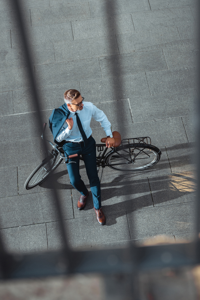vista de ángulo alto de hombre de negocios de mediana edad sosteniendo chaqueta de traje y mirando hacia otro lado mientras está sentado en bicicleta
 - Foto, imagen