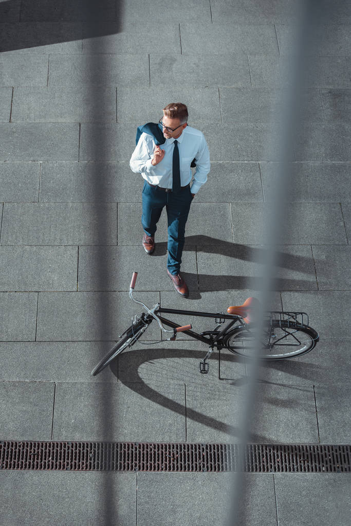 vista de ángulo alto de hombre de negocios con chaqueta de traje y mirando lejos cerca de la bicicleta en la calle
  - Foto, imagen