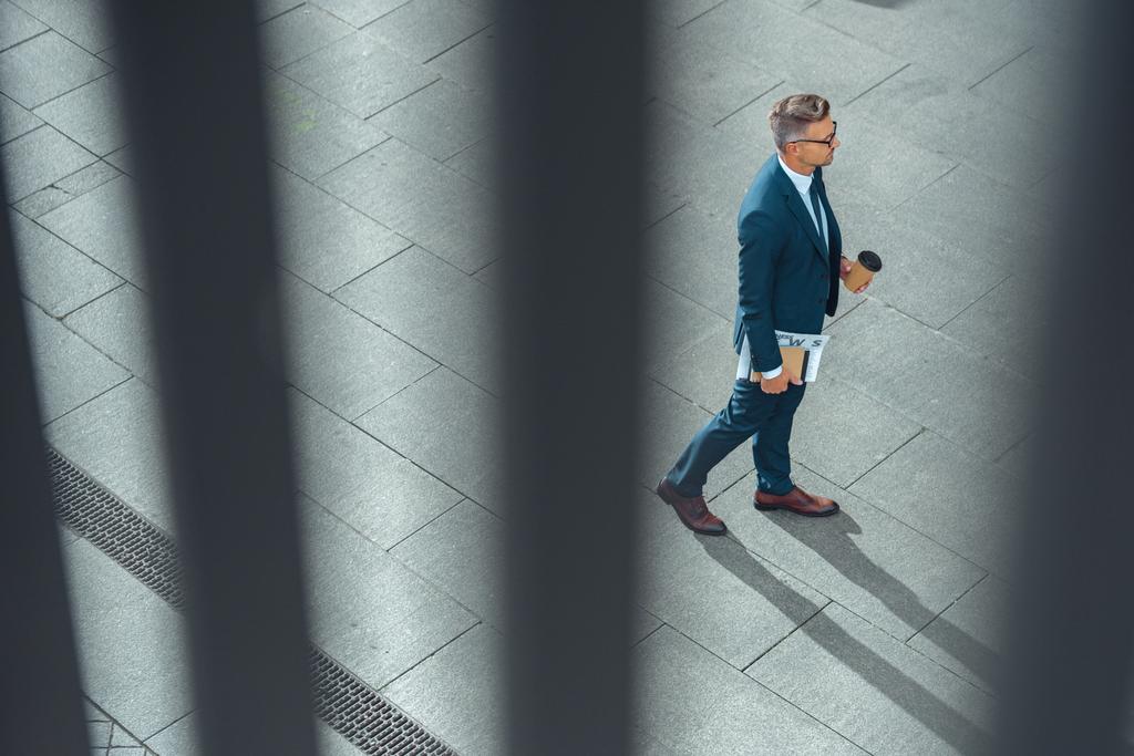 vista de ángulo alto de hombre de negocios con café para ir y periódico caminando por la calle
    - Foto, Imagen