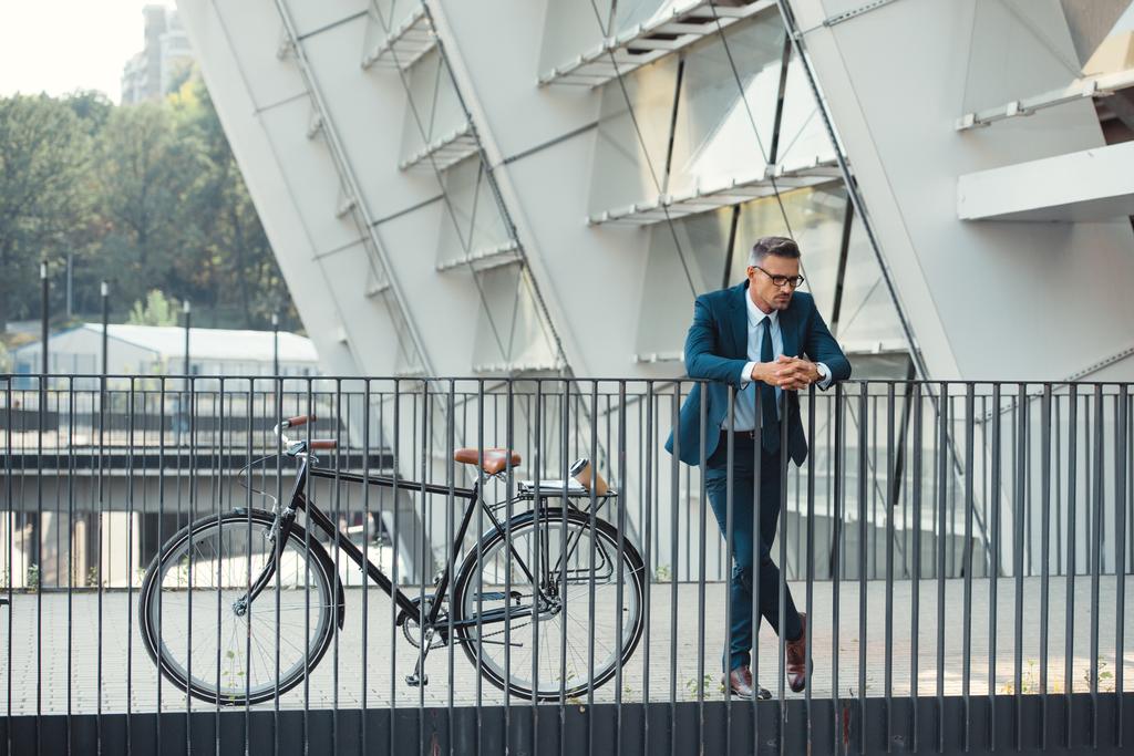 empresário de meia idade bem sucedido em desgaste formal inclinando-se em trilhos perto de bicicleta
 - Foto, Imagem