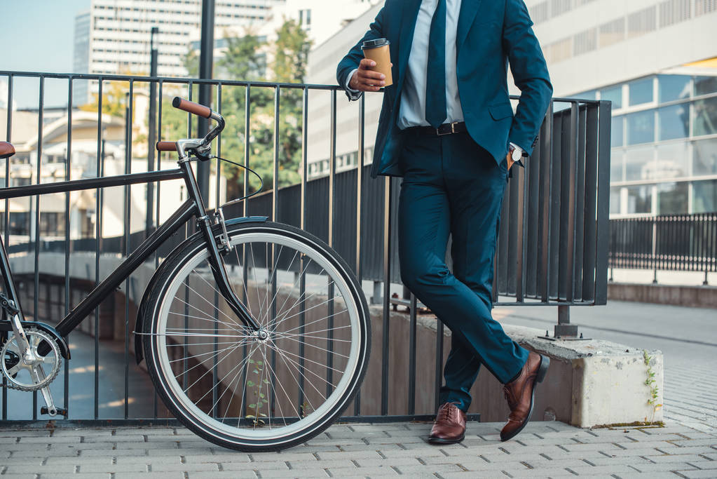 tiro cortado de empresário segurando copo de papel e de pé perto de bicicleta na rua
 - Foto, Imagem