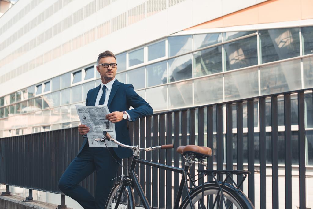 erfolgreicher Geschäftsmann in offizieller Kleidung hält Zeitung in der Hand und schaut weg, während er mit dem Fahrrad auf der Straße steht - Foto, Bild