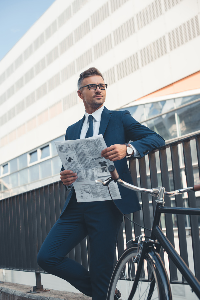 μέσης ηλικίας buisnessman σε κοστούμι και γυαλιά κρατώντας την εφημερίδα και αναζητούν μακριά ενώ στέκεται με ποδήλατο στον δρόμο   - Φωτογραφία, εικόνα