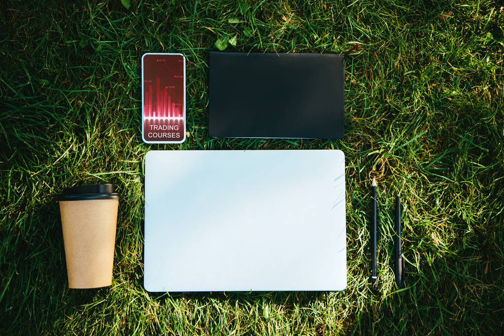 pohled shora na smartphone s obchodní kurzy přístroj a kávu v pohárek na zelené trávě v parku - Fotografie, Obrázek