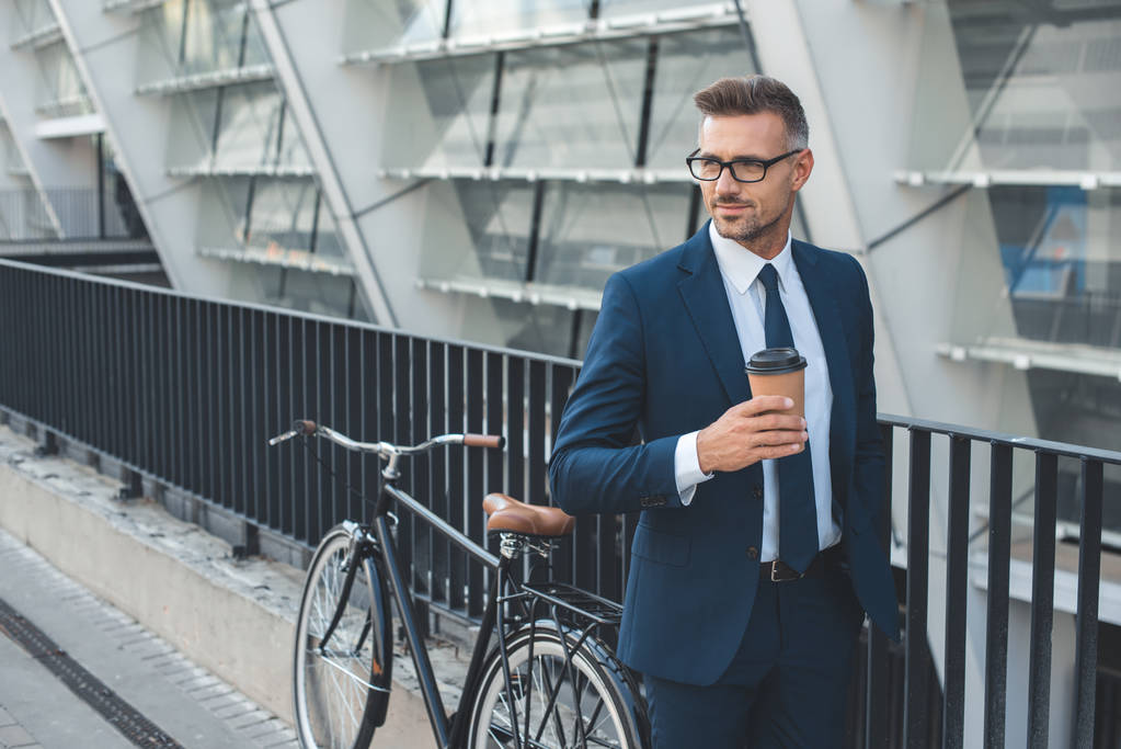 hombre de negocios guapo en traje y gafas con taza de papel y mirando hacia otro lado mientras está de pie con la bicicleta en la calle
 - Foto, Imagen