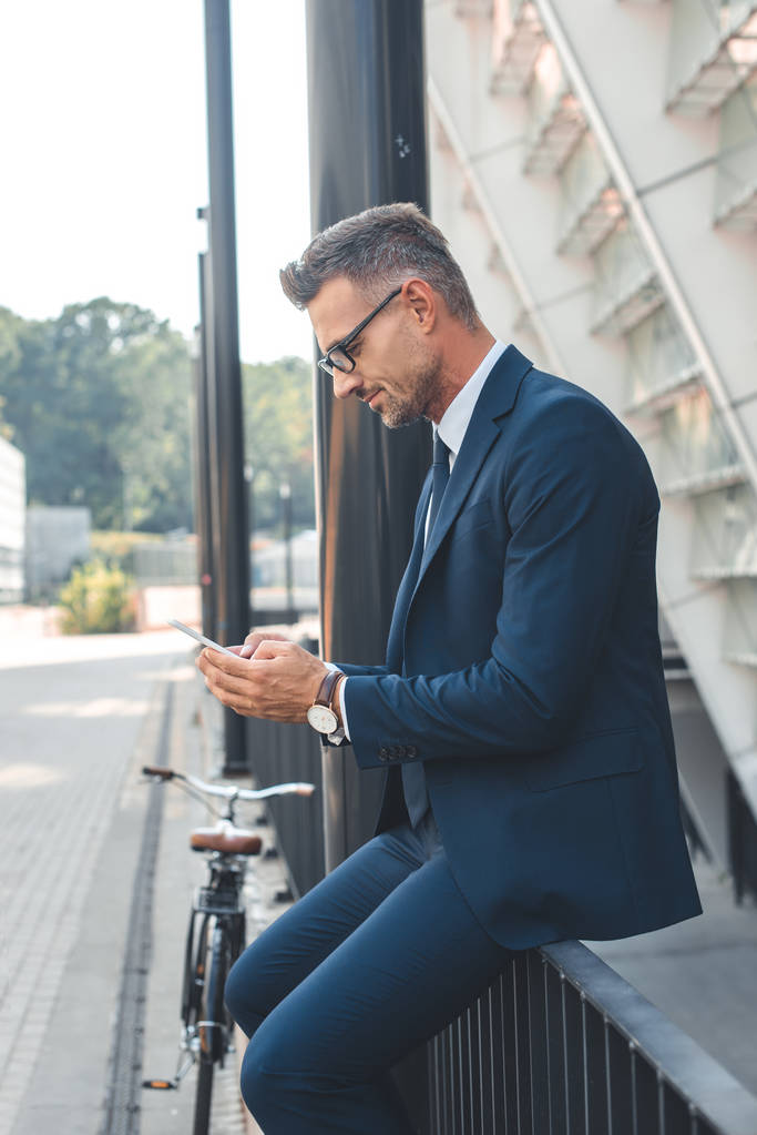 vista laterale dell'uomo d'affari in giacca e cravatta e occhiali utilizzando lo smartphone sulla strada
 - Foto, immagini
