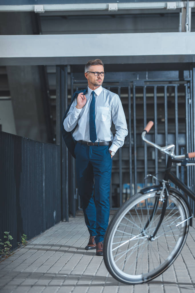 schöner Geschäftsmann mit Brille, Anzugjacke und wegschauendem Blick, während er zu einem auf der Straße geparkten Fahrrad geht   - Foto, Bild
