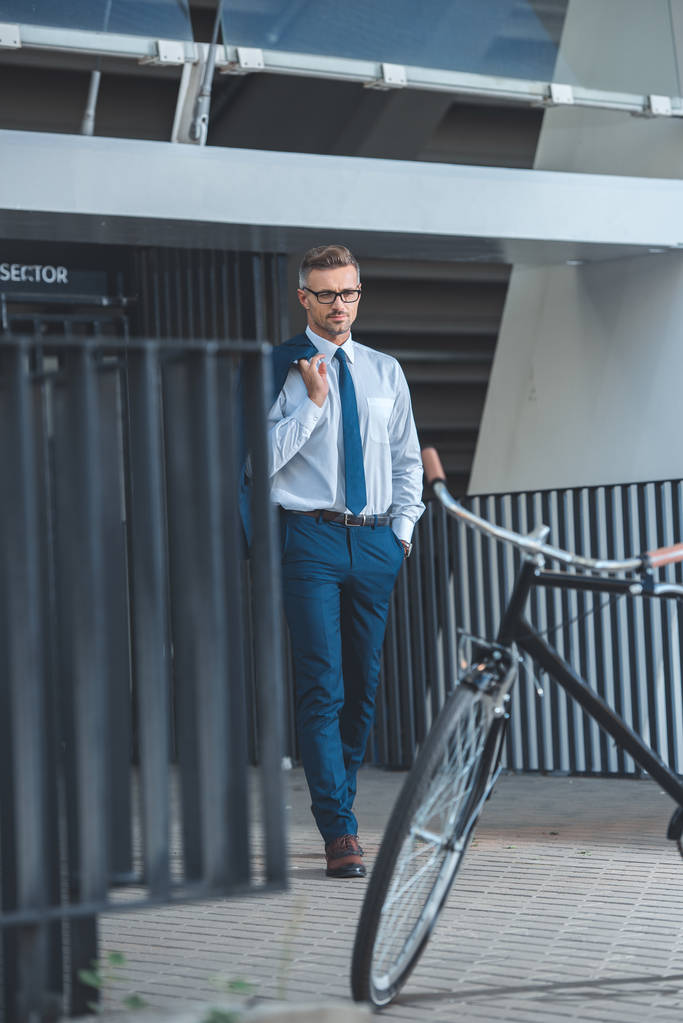 schöner Geschäftsmann mit Brille, Anzugjacke und Fahrrad auf der Straße geparkt   - Foto, Bild