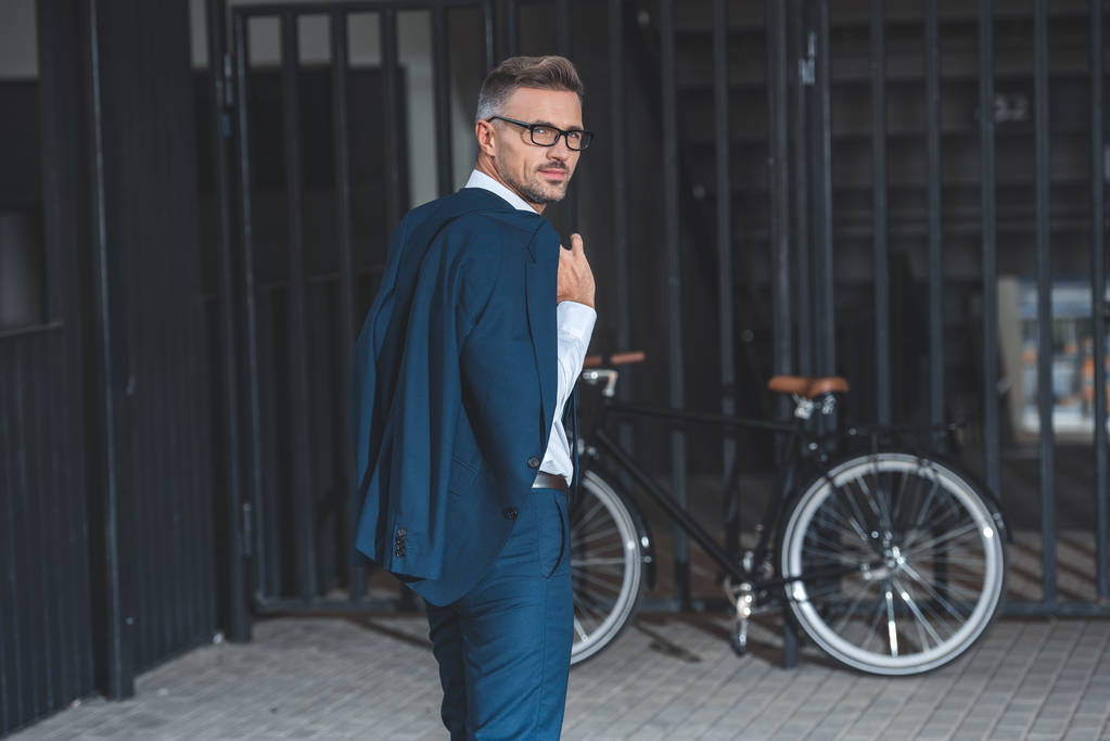 középső éves üzletember a gazdaság szemüveg öltöny zakó és látszó-on fényképezőgép-miközben megy kerékpár parkoló utcában  - Fotó, kép