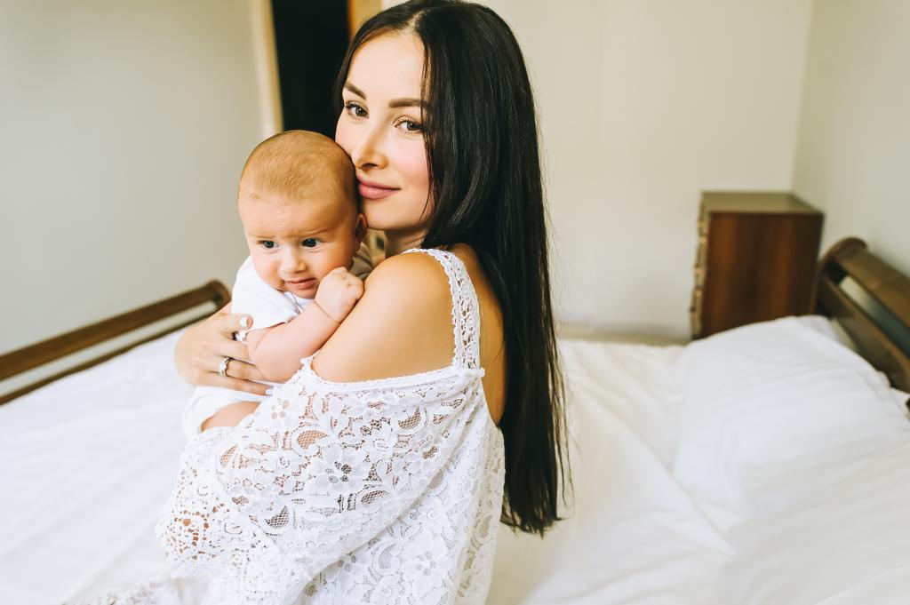 hermosa joven madre con su pequeño bebé sentado en la cama en casa
 - Foto, Imagen