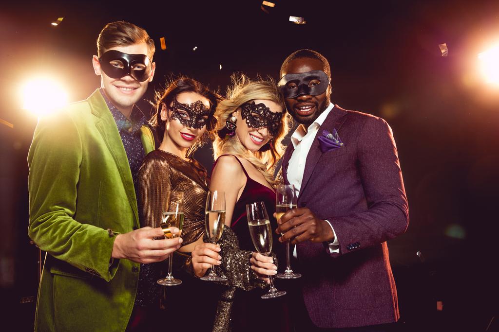 amigos multiétnicos de luxo em máscaras de carnaval celebrando o ano novo com copos de champanhe
 - Foto, Imagem