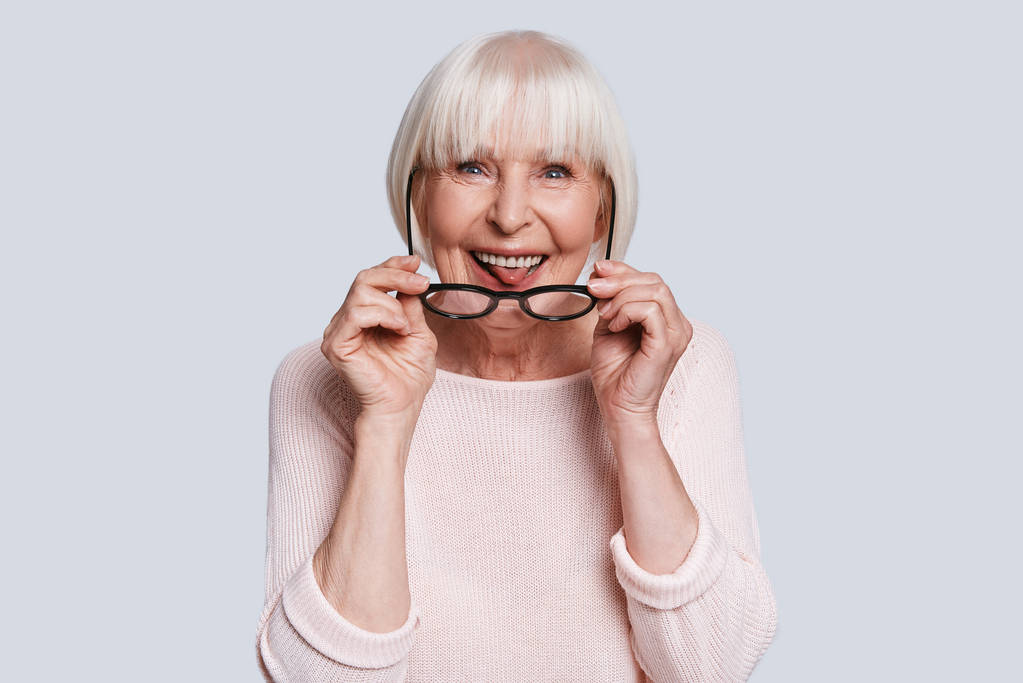 aged woman with short grey hair posing at grey background in studio and adjusting stylish glasses and showing tongue - Photo, Image