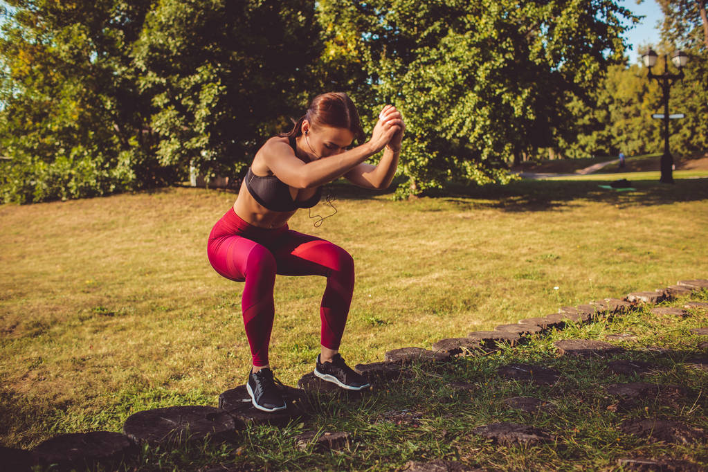 vrouw in rode leggings en sportieve top doen squats in park weide  - Foto, afbeelding