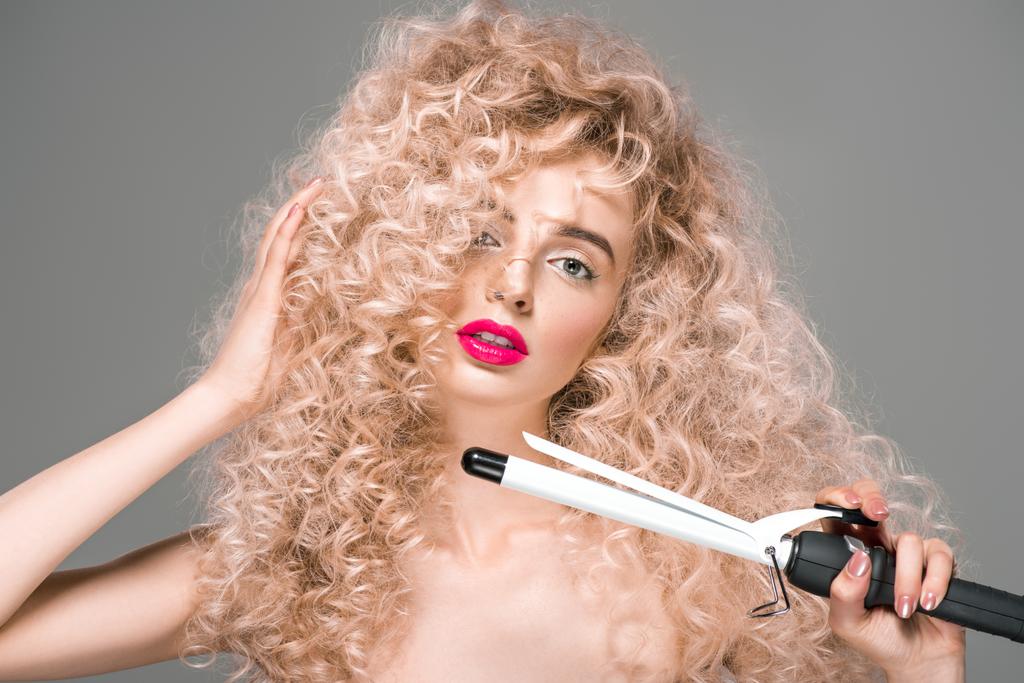 ελκυστική γυμνή κοπέλα με μακριά σγουρά μαλλιά κρατώντας μπικουτί μαλλιά και βλέπουν τα φωτογραφικών μηχανών που απομονώνονται σε γκρι - Φωτογραφία, εικόνα