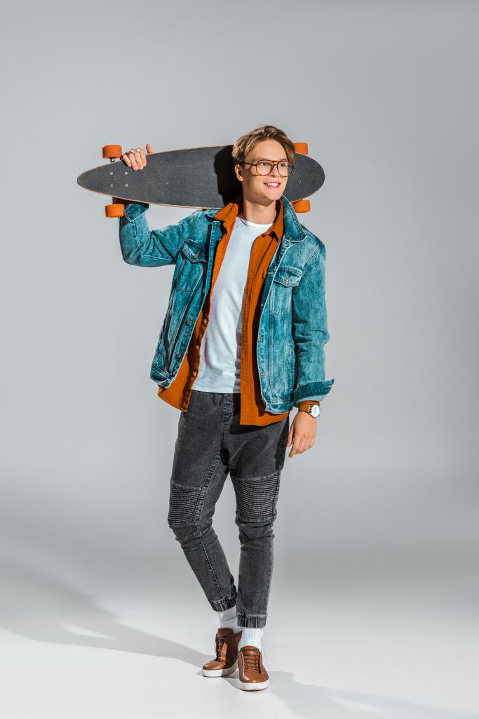 молодий чоловік скейтбордист в джинсовій куртці позує з дошкою на сірому
 - Фото, зображення