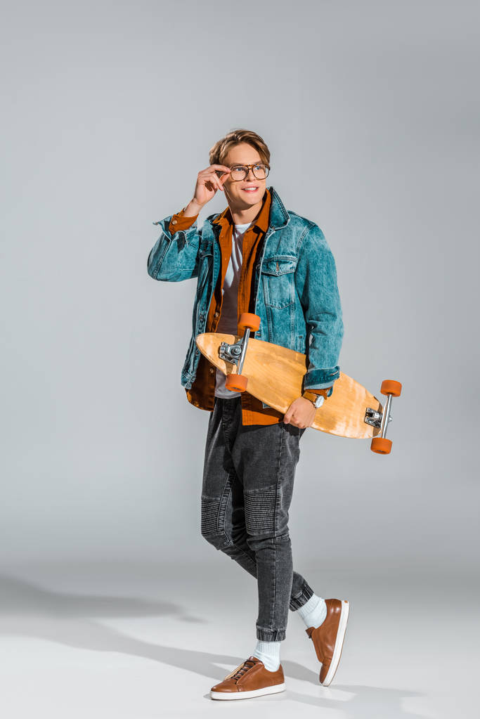 Junger glücklicher Mann in Jeansjacke posiert mit Skateboard auf grau - Foto, Bild