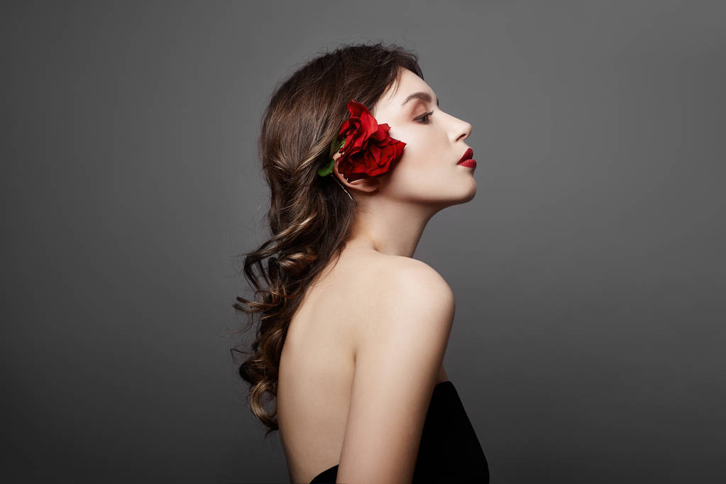 Γυναίκα με ένα μεγάλο κόκκινο λουλούδι στα μαλλιά της. Καστανά μαλλιά κορίτσι με ένα κόκκινο λουλούδι θέτοντας σε γκρι φόντο. Μεγάλα όμορφα μάτια και φυσικό μακιγιάζ. Μακριά σγουρά μαλλιά, τέλειο πρόσωπο - Φωτογραφία, εικόνα