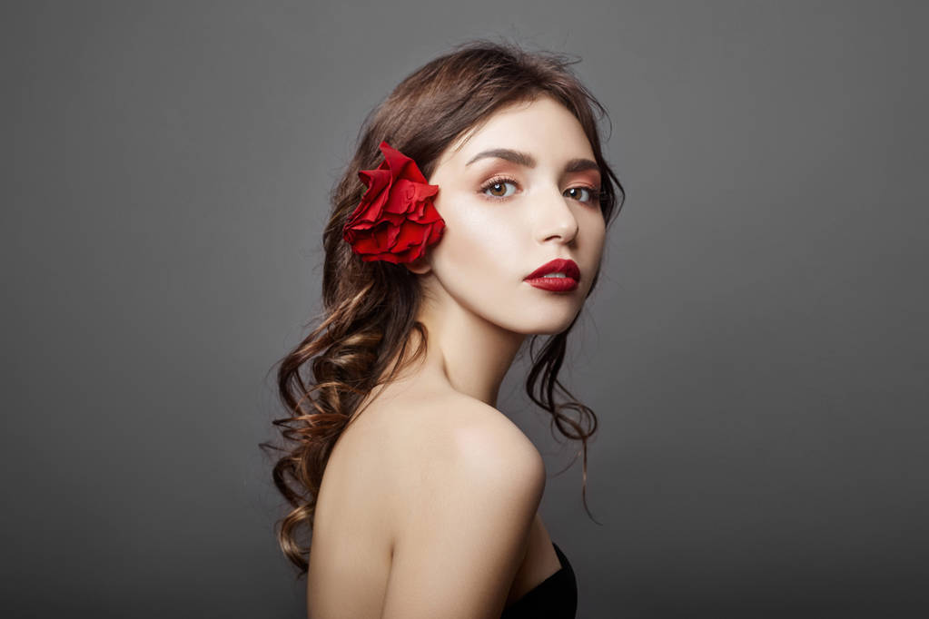 Nő, egy nagy piros virág a hajában. Barna hajú lány pózol egy szürke háttér piros virággal. Nagy gyönyörű szemek és természetes smink. Hosszú göndör hajú, tökéletes arc - Fotó, kép