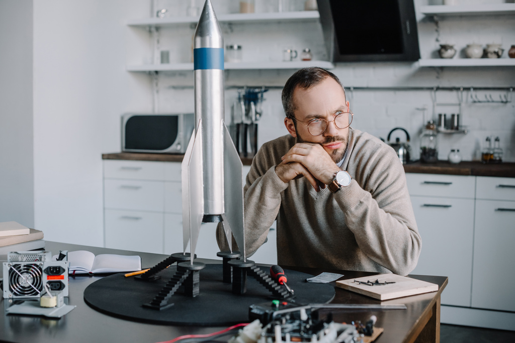 Ingeniero reflexivo sentado en la mesa con el modelo de cohete en casa y mirando hacia otro lado
 - Foto, imagen