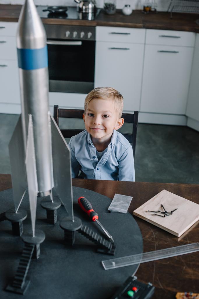 entzückender Junge sitzt am Wochenende mit Raketenmodell in Küche am Tisch - Foto, Bild