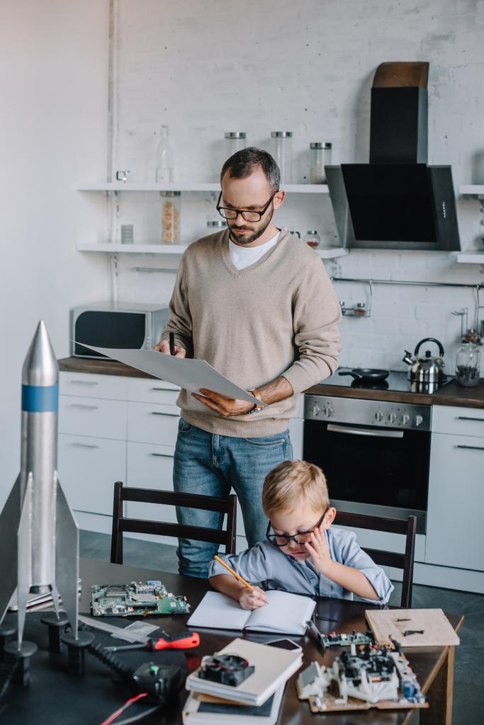 задумчивый отец смотрит на план и сын писать что-то в тетради дома
 - Фото, изображение