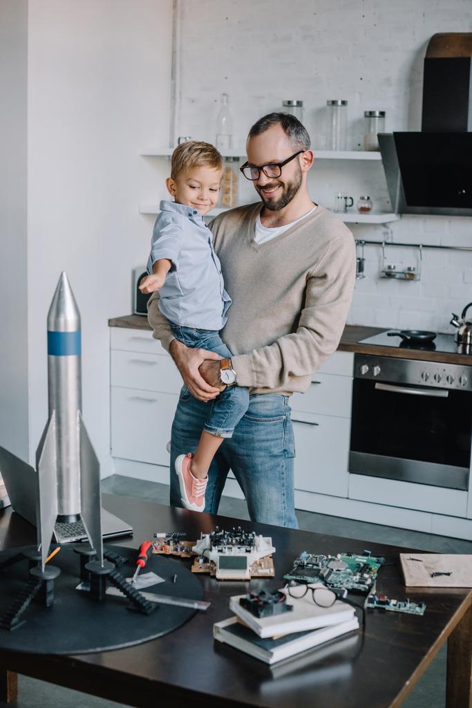 smiling father carrying son and looking at rocket model at home - Photo, Image