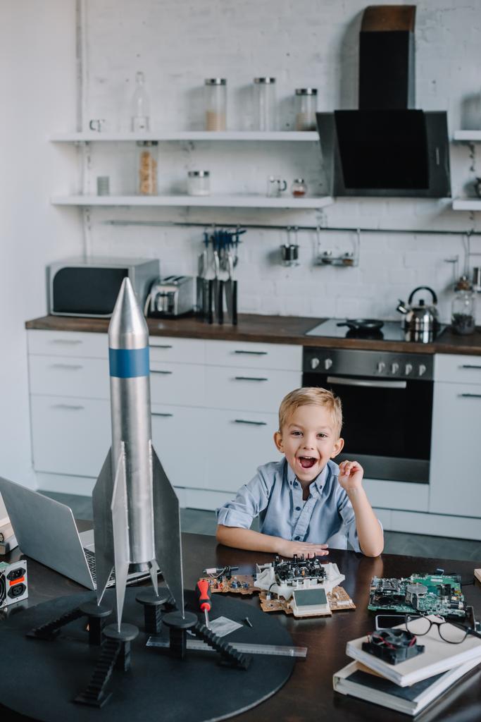 szczęśliwy chłopiec siedzi przy stole z model rocket w kuchni i patrząc na kamery - Zdjęcie, obraz