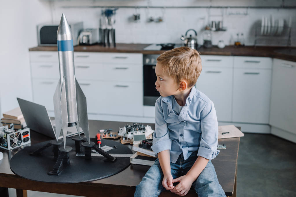 podrážděný grimasy chlapce, který seděl na stole u modelu rakety v kuchyni na víkend - Fotografie, Obrázek