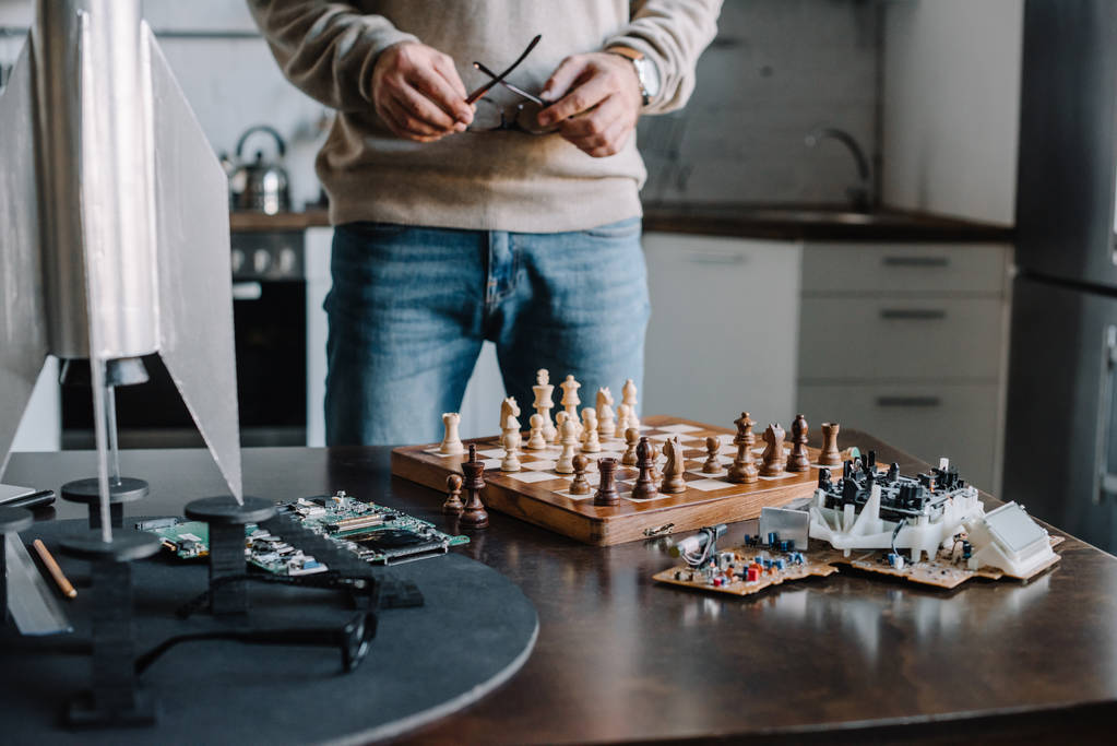 imagen recortada del hombre de pie cerca de la mesa con tablero de ajedrez y la celebración de gafas en casa
 - Foto, imagen