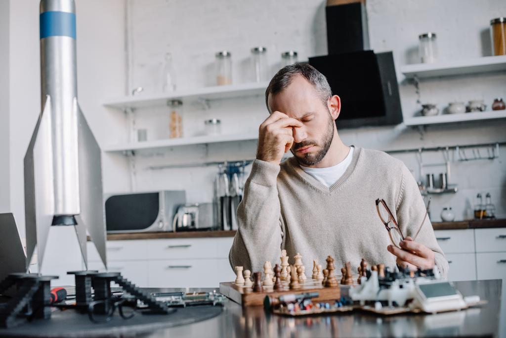 zmęczony przystojny mężczyzna gra w szachy i dotykając nosa w domu - Zdjęcie, obraz