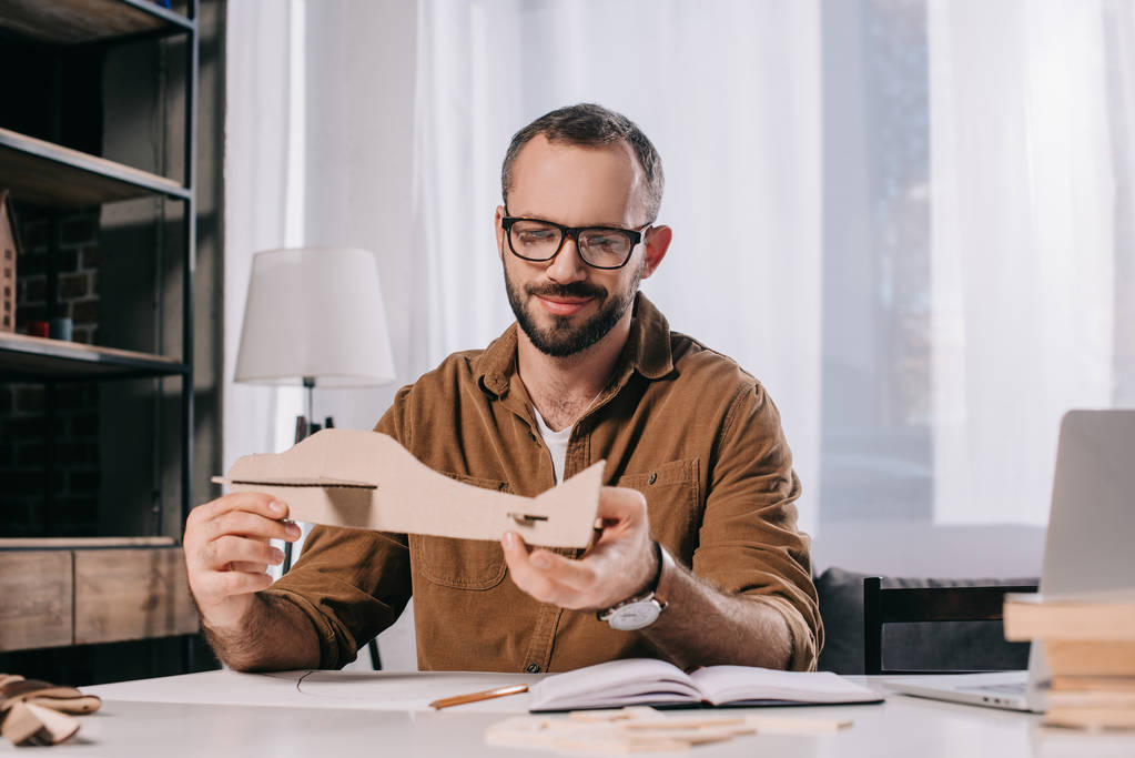 όμορφος χαμογελαστός άνθρωπος σε γυαλιά κρατώντας χάρτινο αεροπλάνο ενώ μοντελοποίηση στο σπίτι  - Φωτογραφία, εικόνα